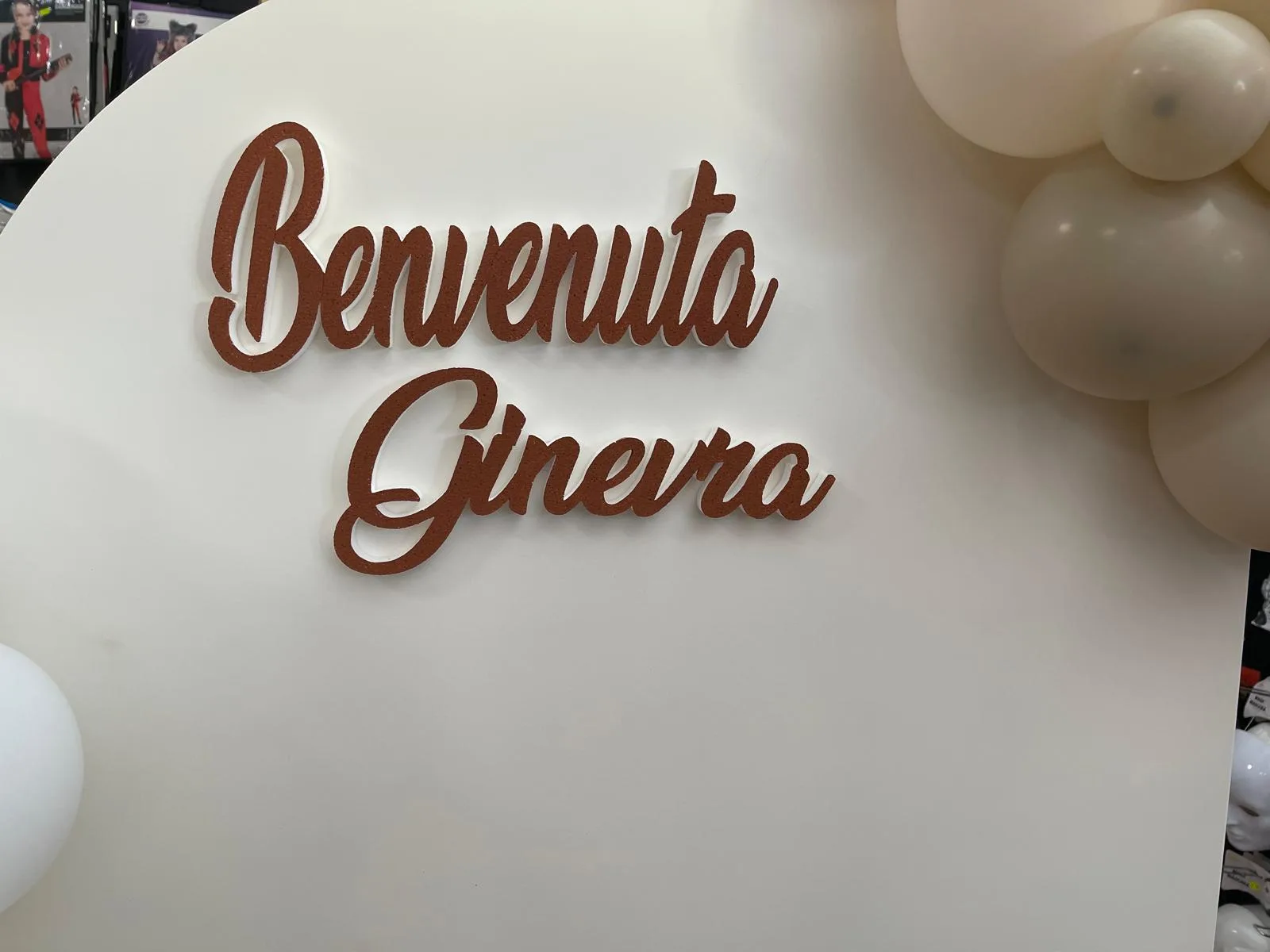 Scritta in corsivo 'Benvenuta Ginevra' tagliata nel polistirolo color terracotta, applicata su un pannello bianco accanto a palloncini avorio