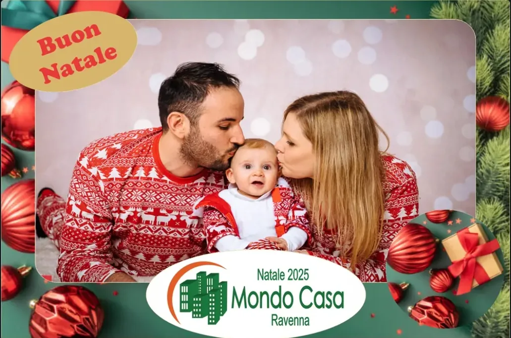 Cornice natalizia con il logo di Mondo Casa Ravenna: mamma e papà in maglione rosso con le renne baciano il loro bambino, tra palline rosse e rami di abete