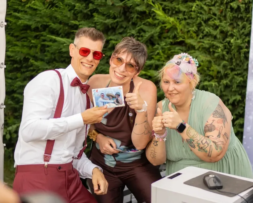 Tre invitati sorridenti in giardino mostrano la foto appena stampata dalla cabina, con il pollice alzato accanto alla stampante