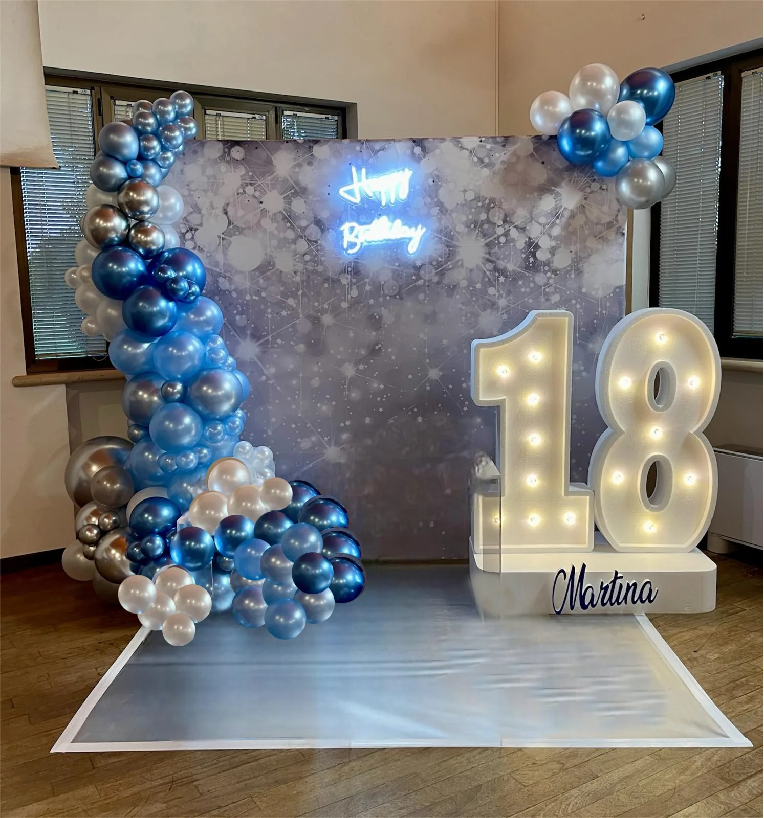 Postazione per un diciottesimo in sala: numero 18 luminoso con il nome Martina, colonna di palloncini blu e argento, insegna al neon Happy Birthday e tavolino con la torta e la bottiglia