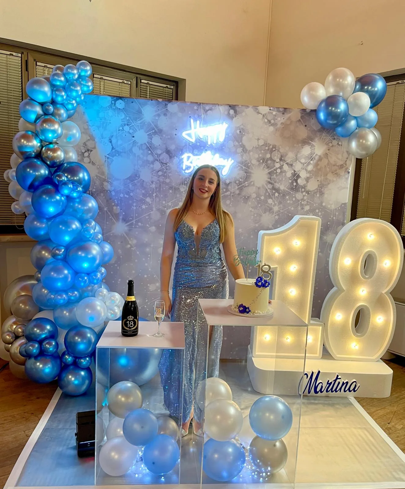 Fondale argento con insegna al neon Happy Birthday, ghirlanda di palloncini blu e cromati e numero 18 luminoso su base con il nome Martina