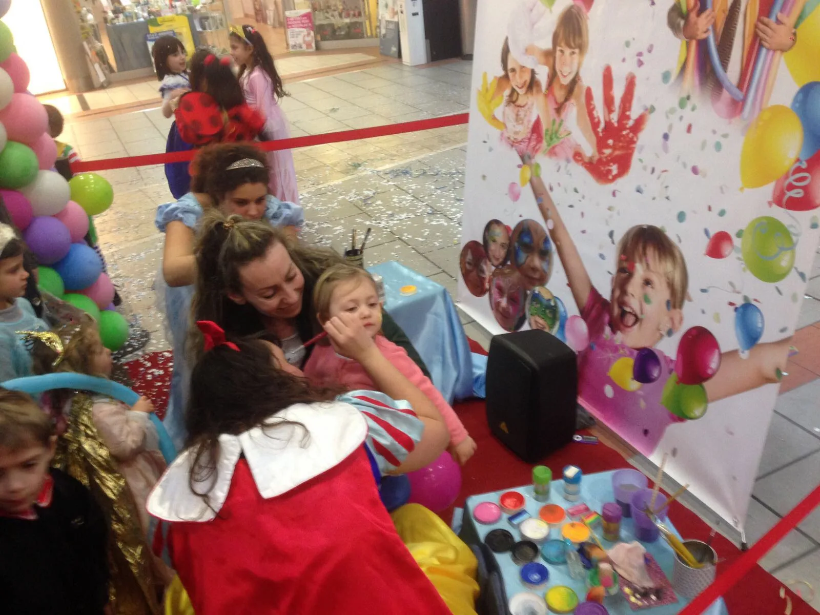 Truccatrice che dipinge il viso di una bambina alla postazione trucchi in una galleria, con i colori sul tavolino e il pannello colorato alle spalle