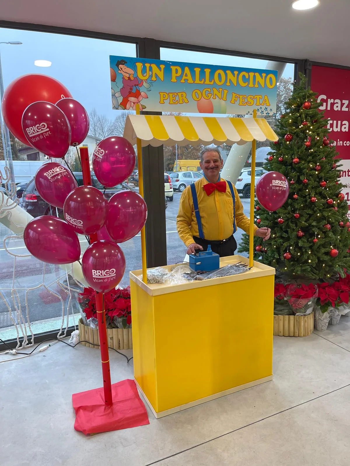 Banchetto giallo con tettoia a righe, gonfiatore e insegna 'Un palloncino per ogni festa', con albero porta-palloncini rossi personalizzati all'ingresso di un negozio