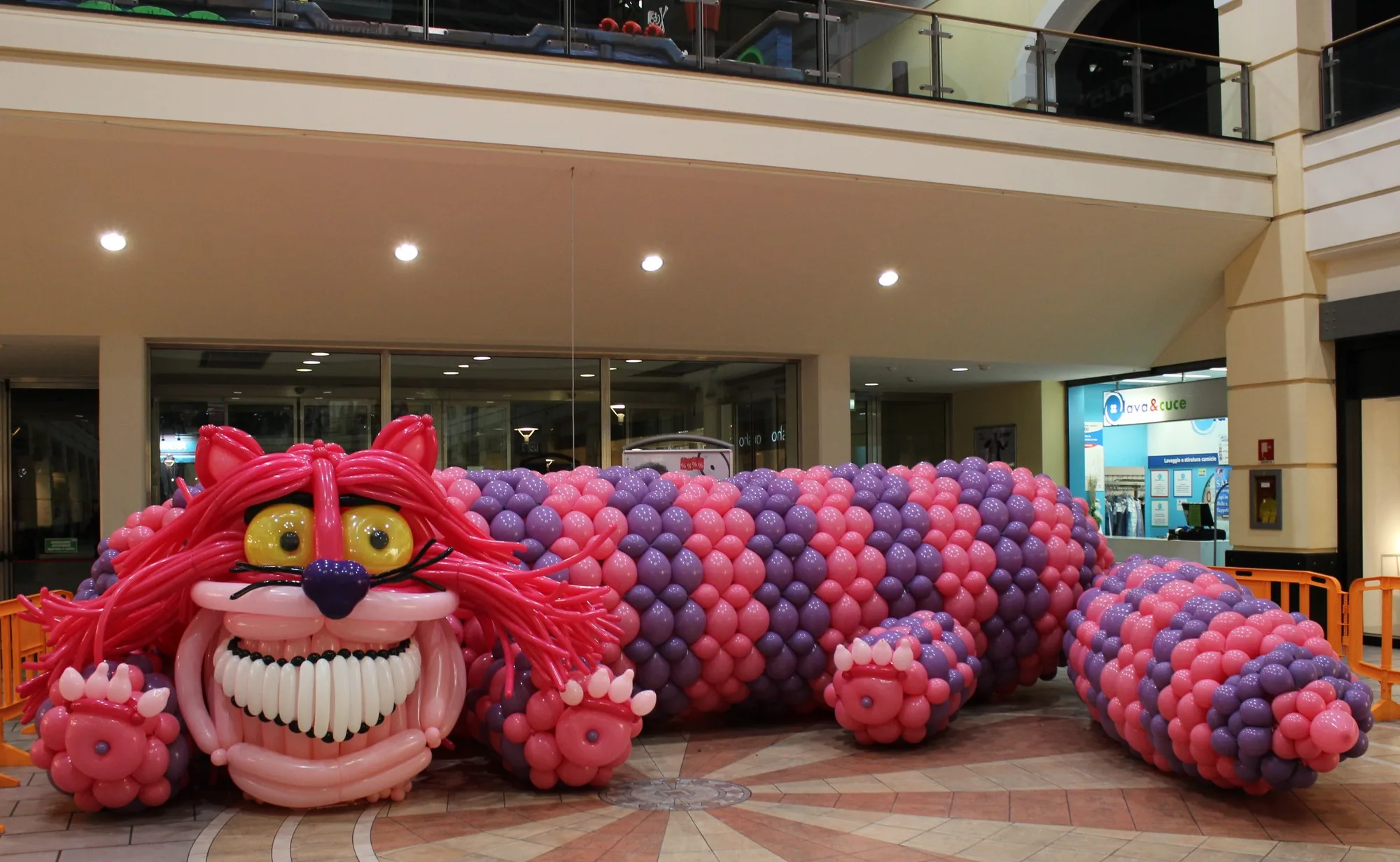 Gatto gigante di palloncini rosa, fucsia e viola lungo diversi metri, allestito nella galleria di un centro commerciale