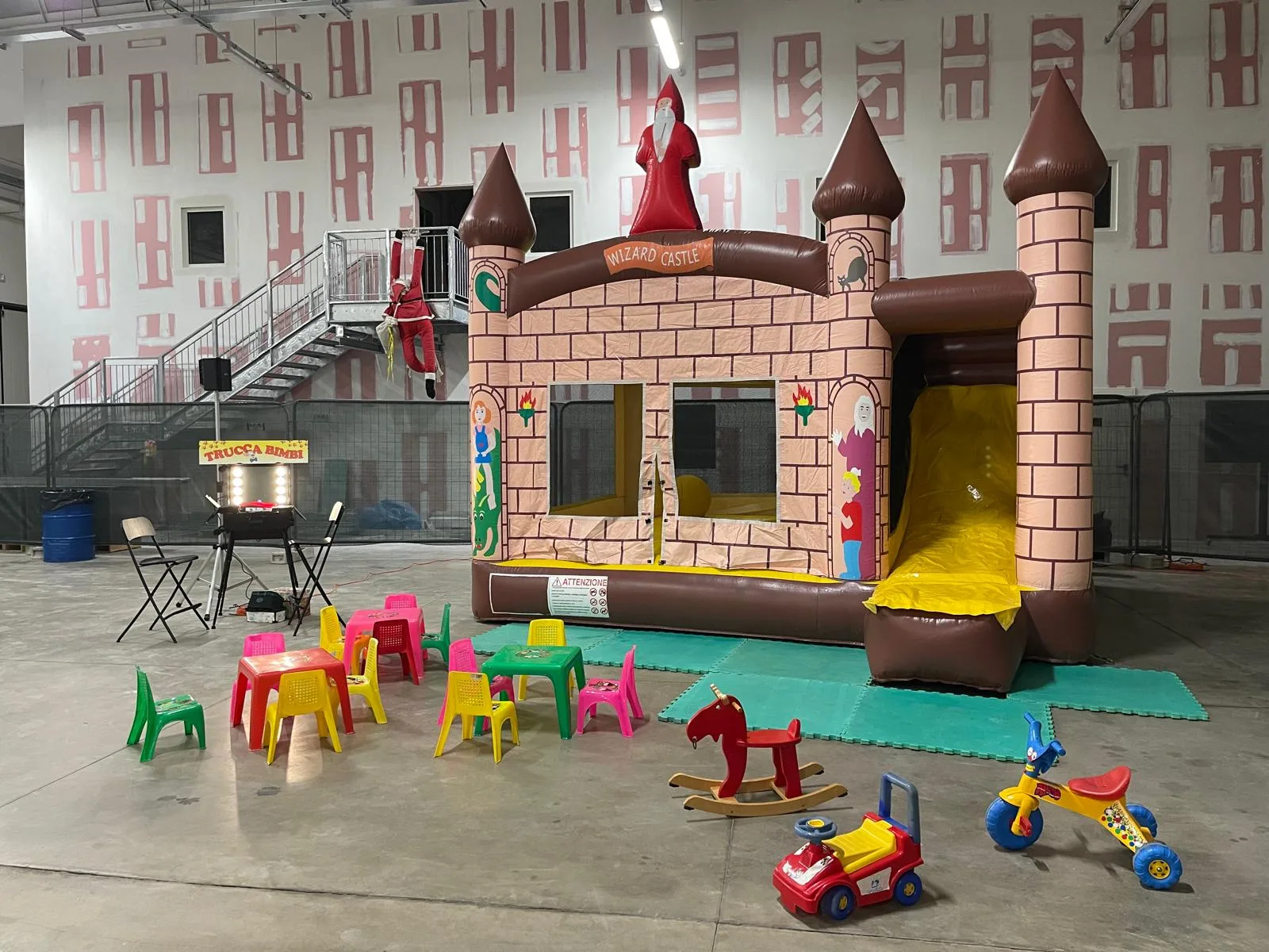 Allestimento del baby parking in un capannone: tavolini e seggioline di plastica colorata su tappeti morbidi, cavallo a dondolo, tricicli, postazione trucca bimbi e castello gonfiabile con scivolo