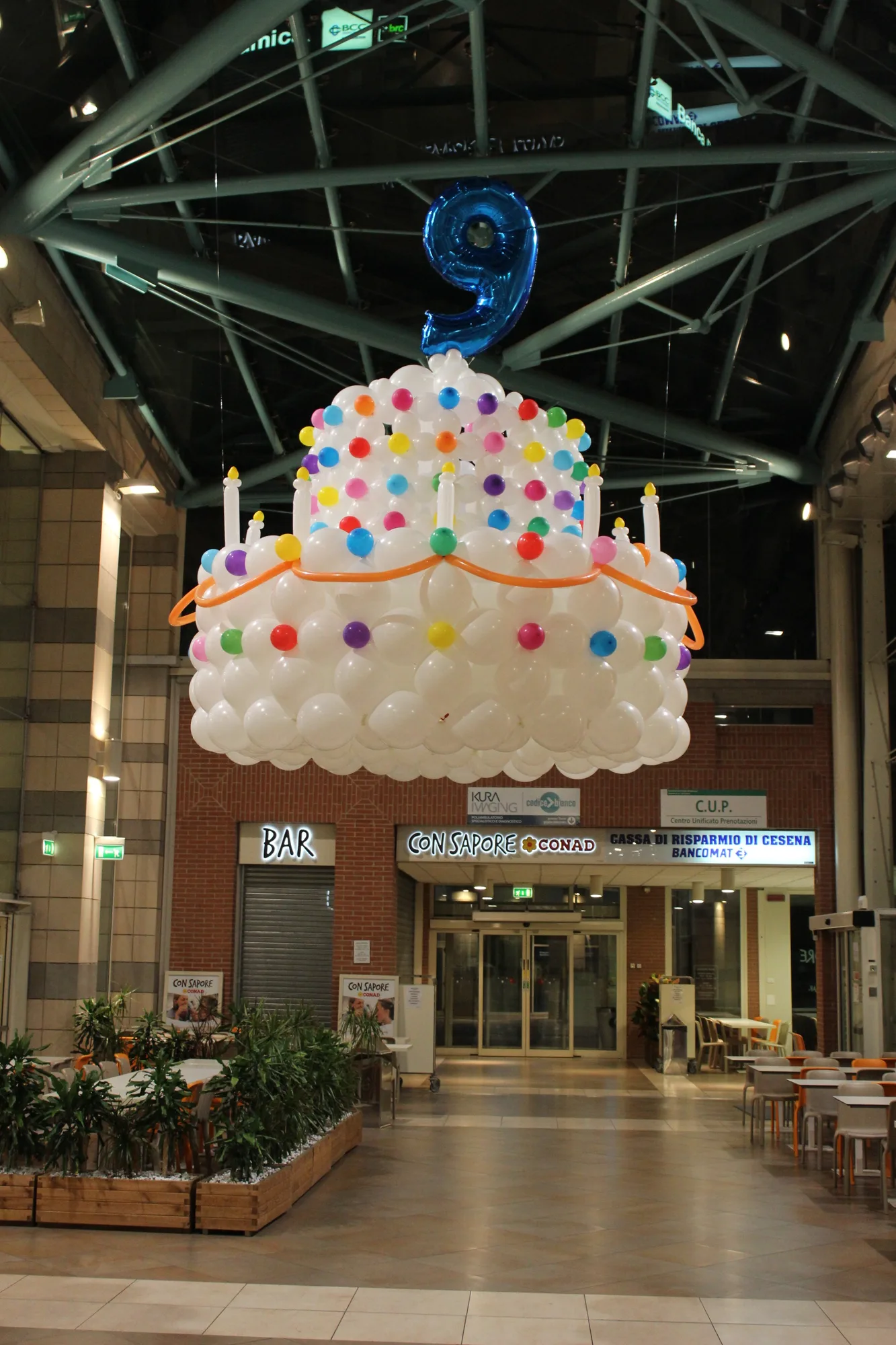 Torta di palloncini a tre piani bianca, azzurra e rossa più alta di una persona, con candeline blu e numero 25 in mylar sulla cima