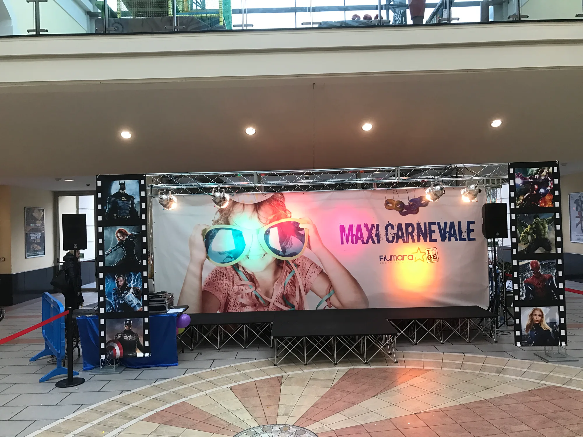 Traliccio in alluminio con fari accesi che regge un grande fondale stampato di Carnevale sopra un palco nero, in una galleria commerciale