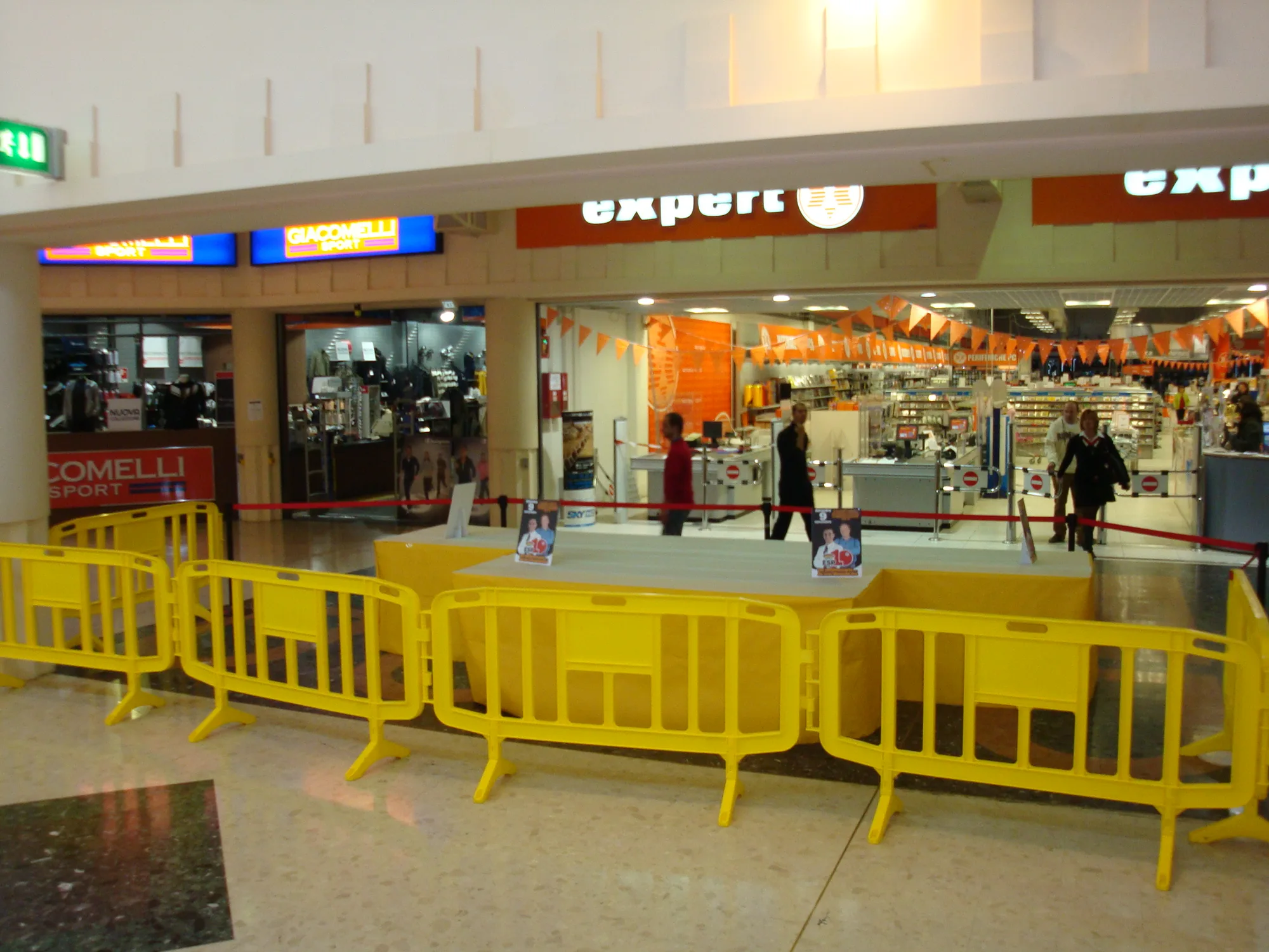 Fila di transenne gialle che delimita un palco allestito davanti ai negozi di un centro commerciale