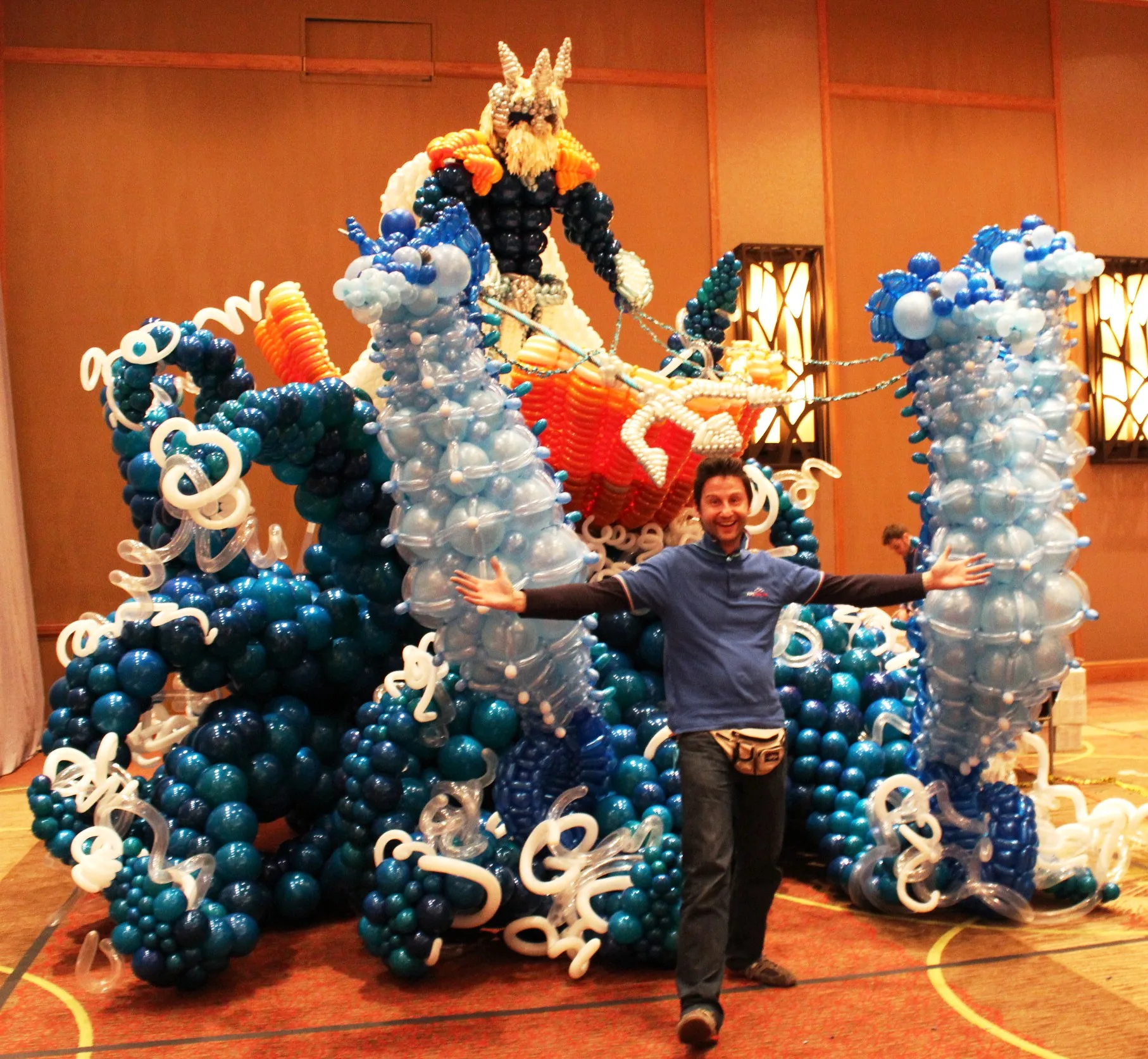 Scenografia gigante di palloncini blu e azzurri con onde, spuma e un tritone in cima a una nave, con davanti una persona a braccia aperte che ne mostra la dimensione