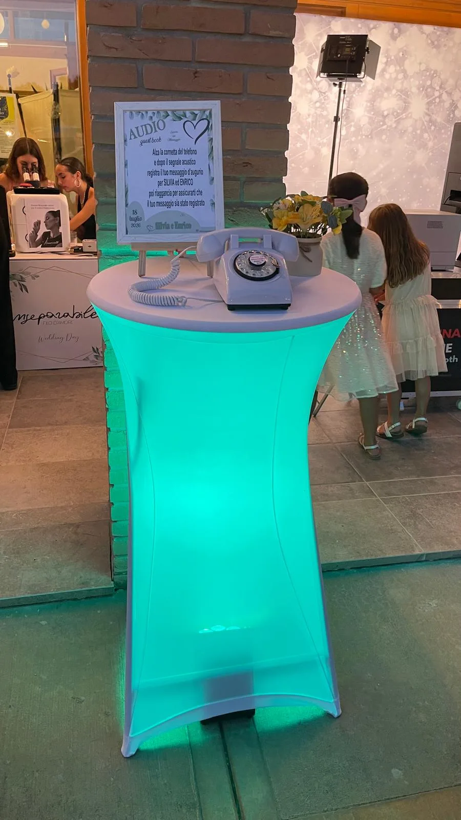 La postazione dell'audio guest book di sera, con il tavolo alto illuminato di verde acqua, il telefono bianco e il cartello con i nomi degli sposi