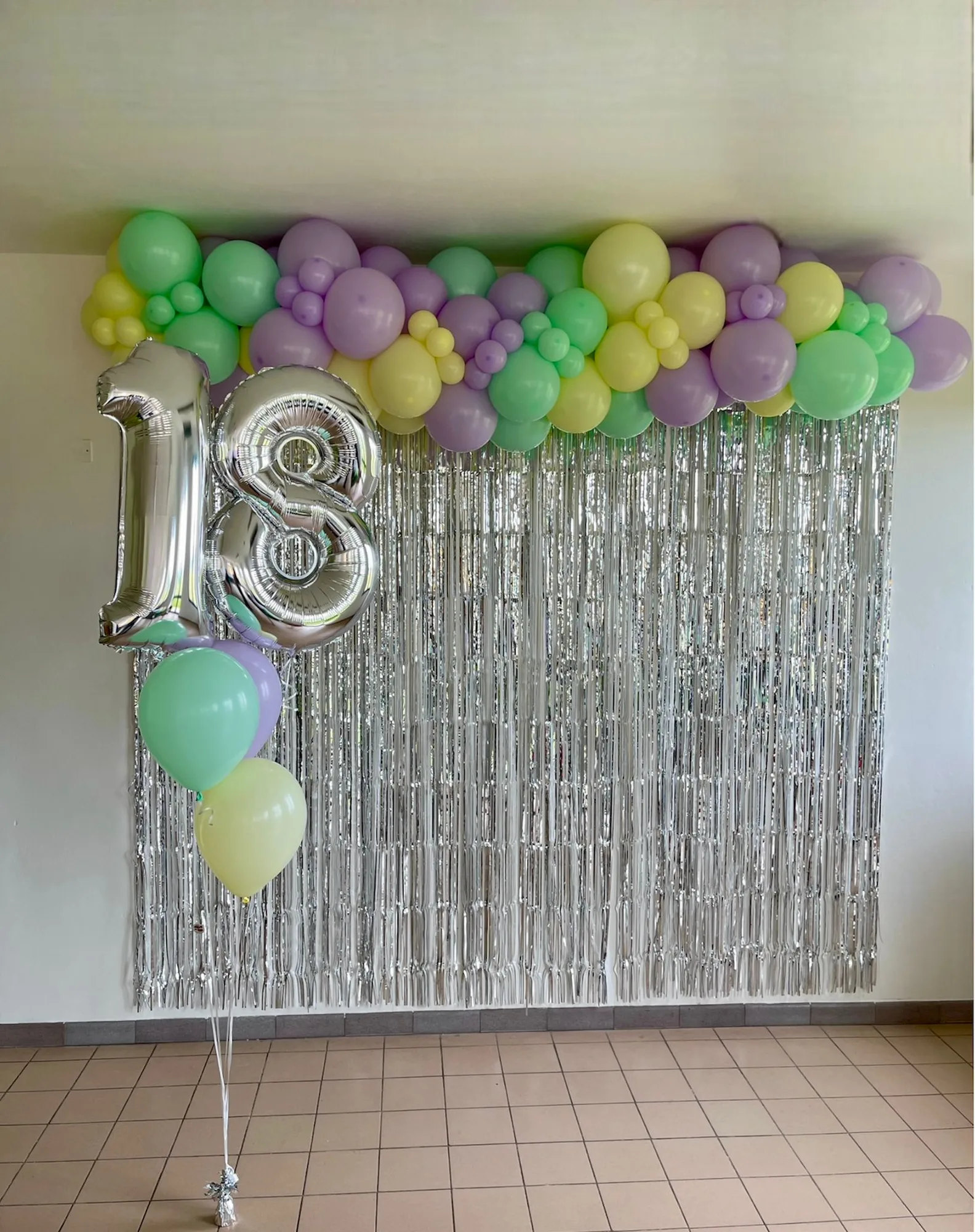 Parete con tenda a frange argento, ghirlanda di palloncini verde menta, lilla e giallo pastello e numero 18 in mylar argento