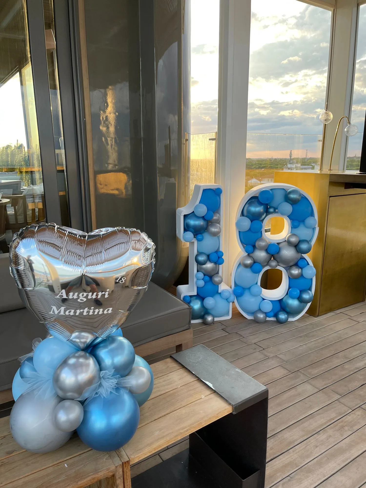 Numero 18 sagomato riempito di palloncini azzurri, blu e argento su una terrazza al tramonto, con cuore in mylar stampato Auguri Martina su base di palloncini