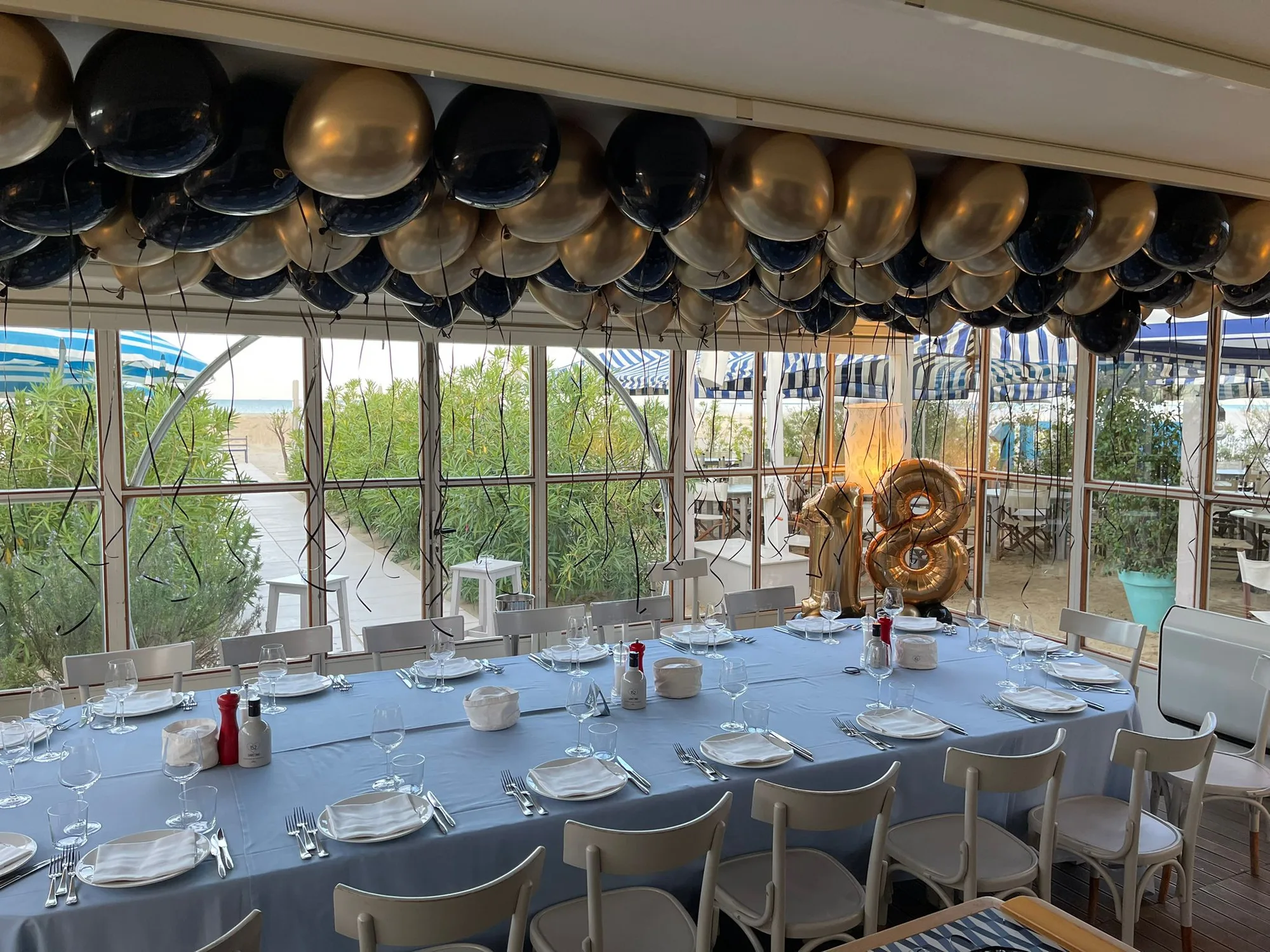 Soffitto di uno stabilimento balneare coperto di palloncini a elio neri e oro con i nastri lunghi sopra la tavolata e numero 18 dorato in fondo