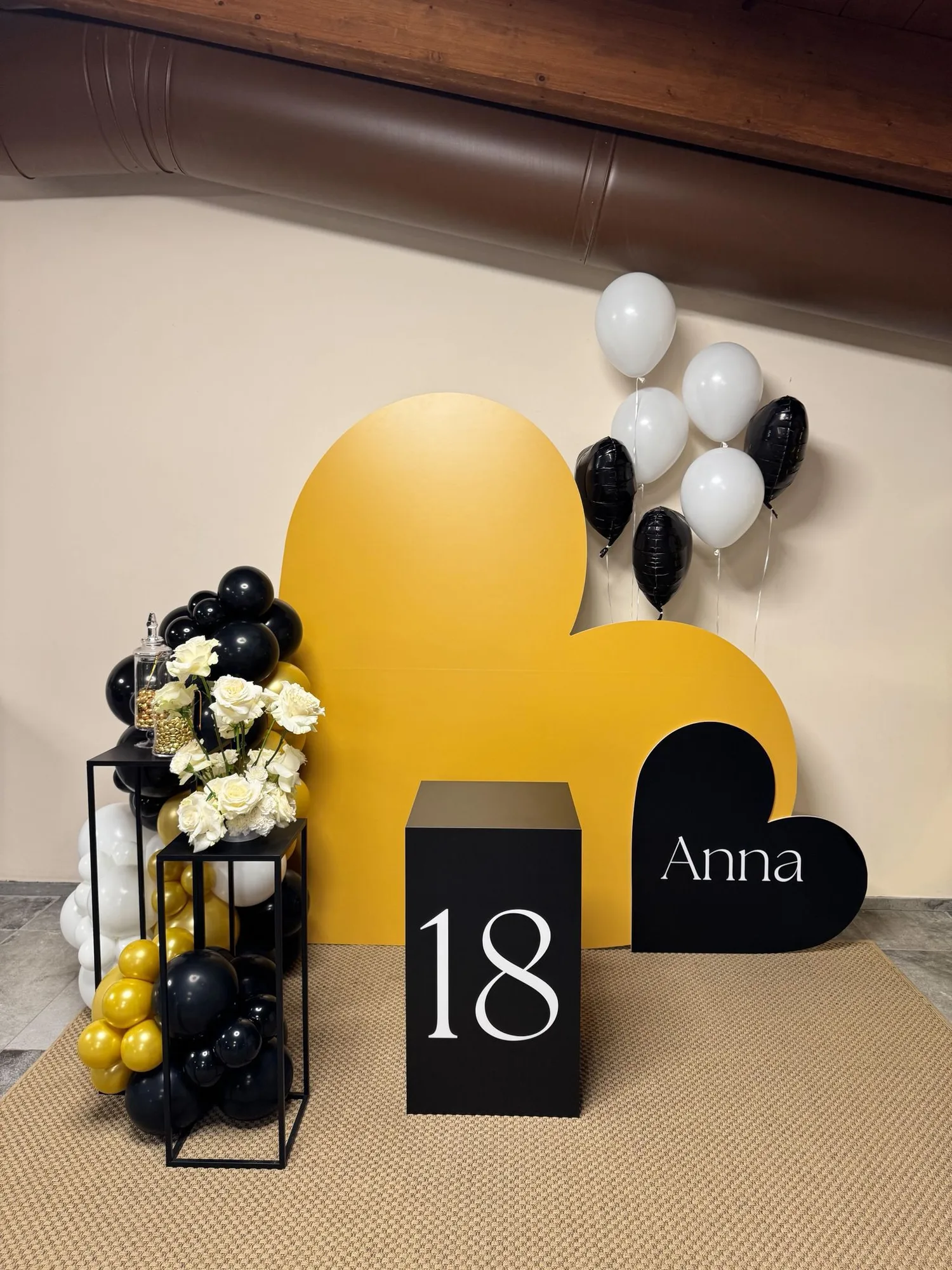 Fondale a nuvola giallo senape con cubo nero e numero 18, cuore nero con il nome Anna, palloncini bianchi e neri e composizione di rose bianche