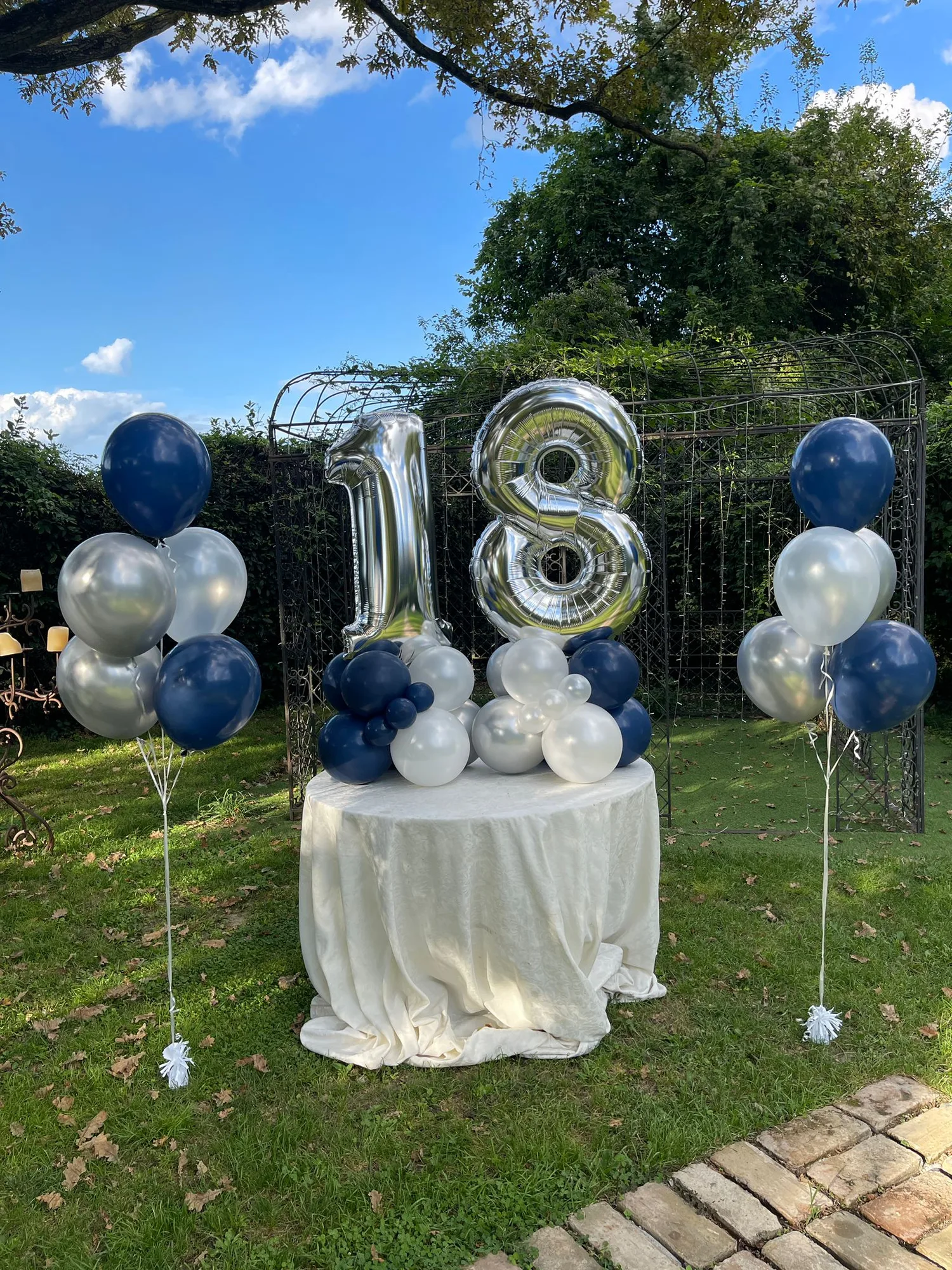 Tavolo tondo in giardino con numero 18 in mylar argento, base di palloncini blu notte e bianchi e due mazzi a elio ai lati