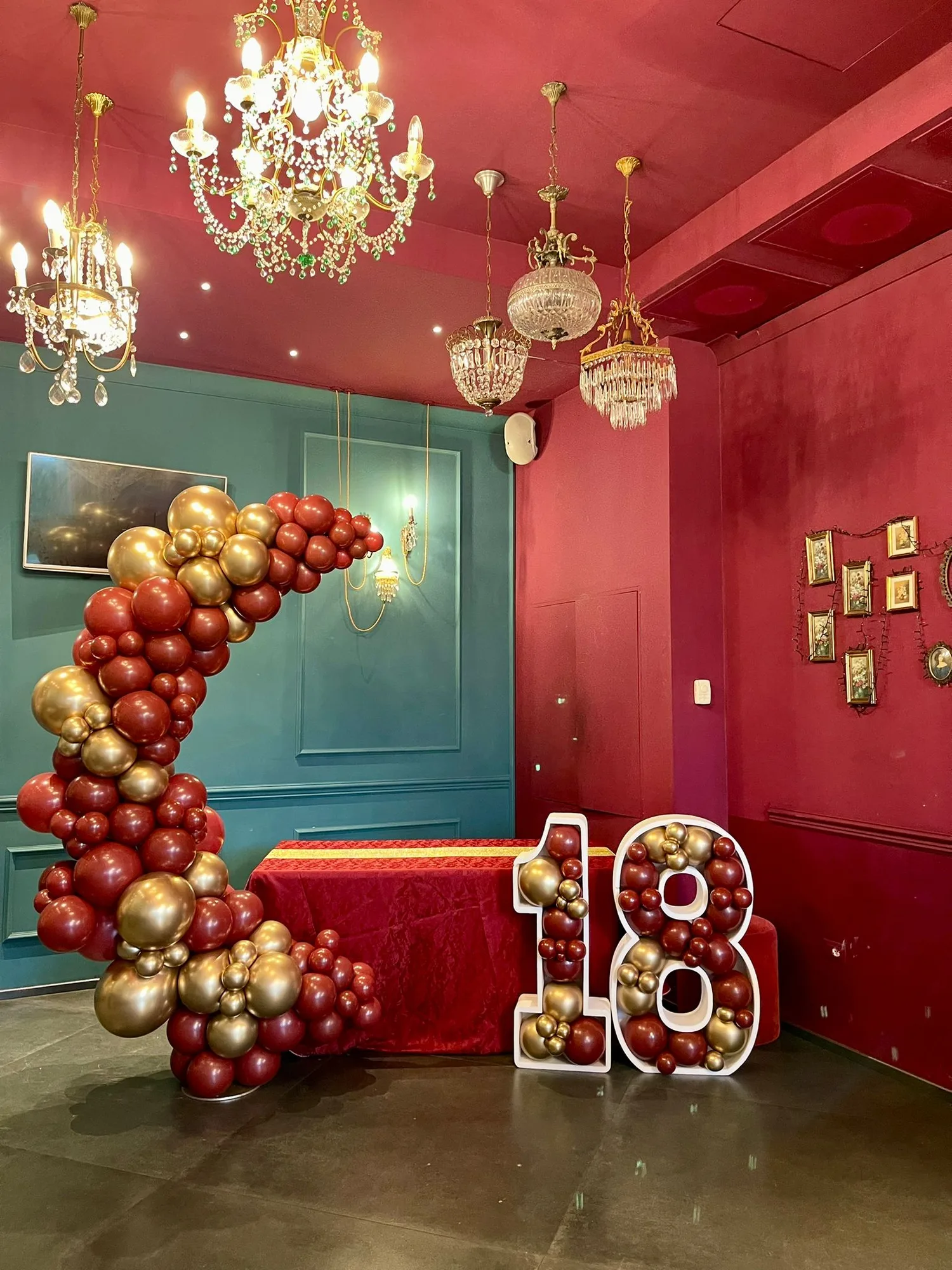 Sala storica con pareti rosse e verdi: cerchio di palloncini rosso scuro e oro cromato accanto al tavolo drappeggiato e numero 18 sagomato pieno di palloncini