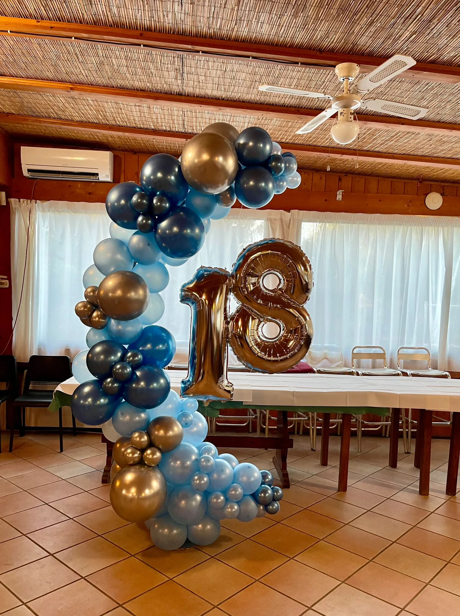 Cerchio di palloncini azzurri, blu e oro cromato con numero 18 in mylar oro davanti alla tavolata di una sala per feste