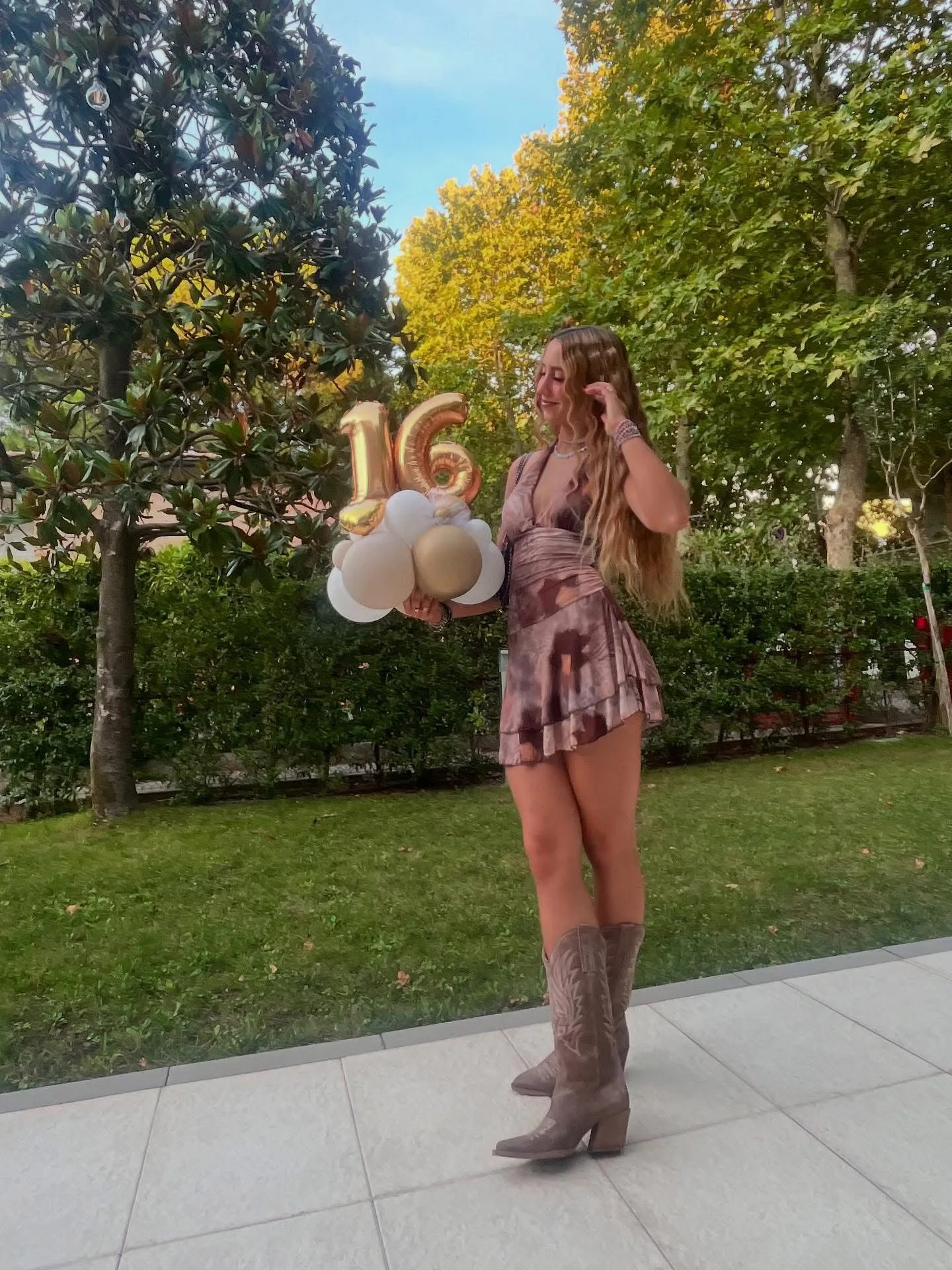 Ragazza in giardino con un mazzo di palloncini a elio bianchi e oro sormontato dal numero 16 in mylar
