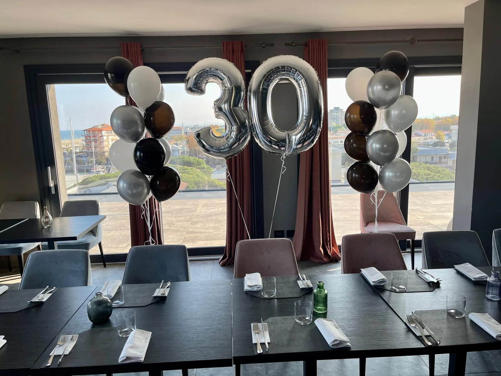 Numero 30 in mylar argento e due colonne di palloncini bianchi, neri e argento allestiti in una sala ristorante con vista sul porto