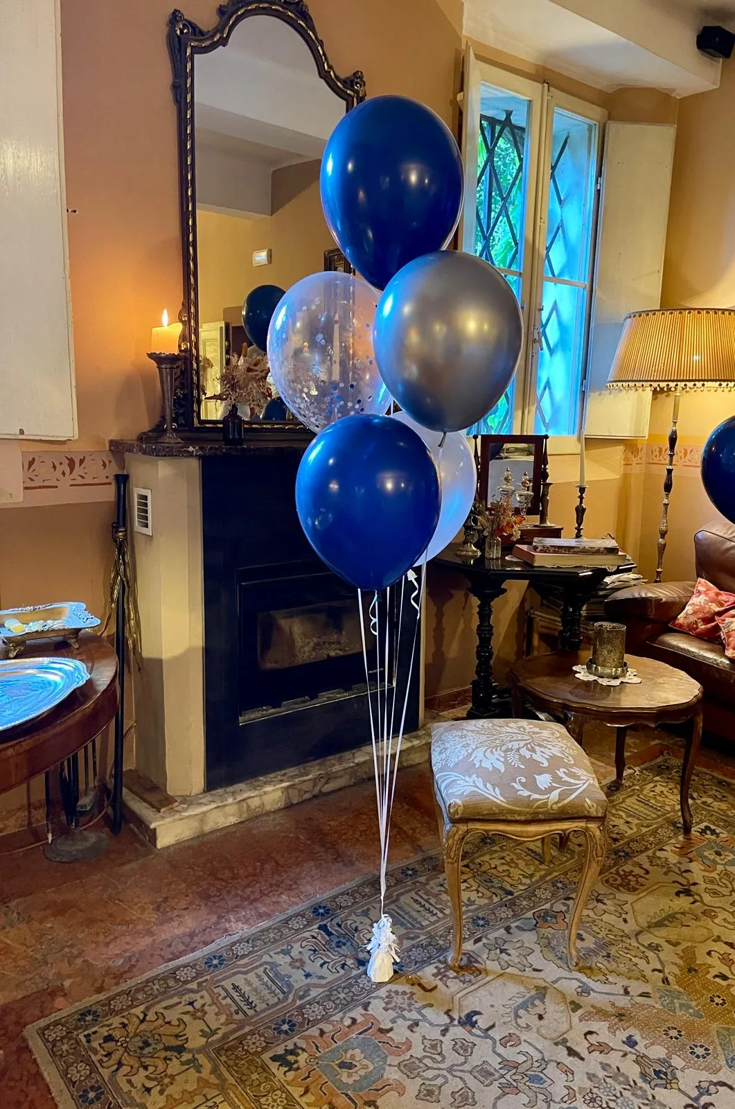 Mazzo di palloncini a elio blu, argento e trasparenti con coriandoli, appoggiato al camino di un salotto