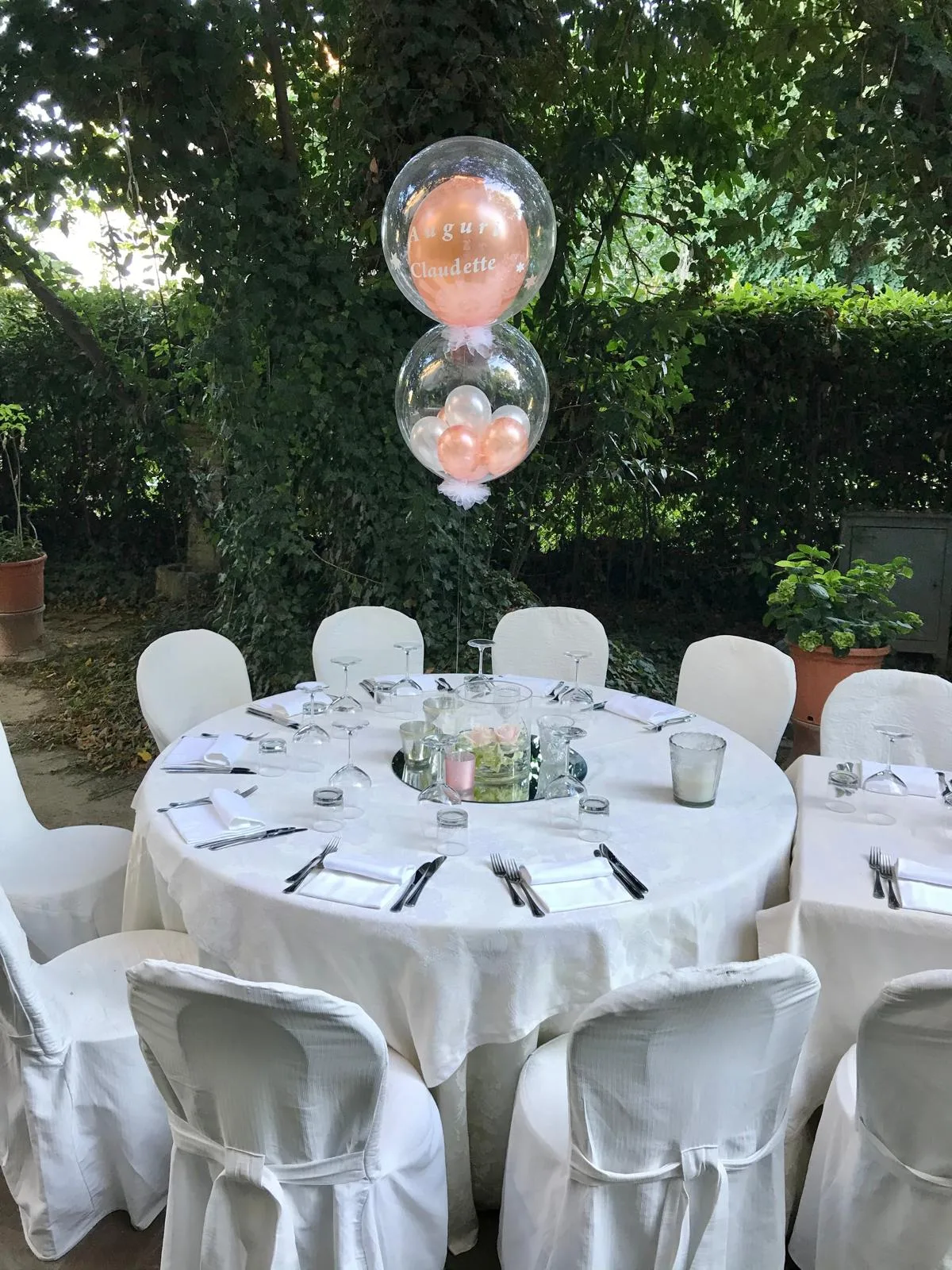 Centrotavola con due palloncini bubble trasparenti, uno con la scritta Auguri Claudette, sopra un tavolo rotondo apparecchiato in giardino