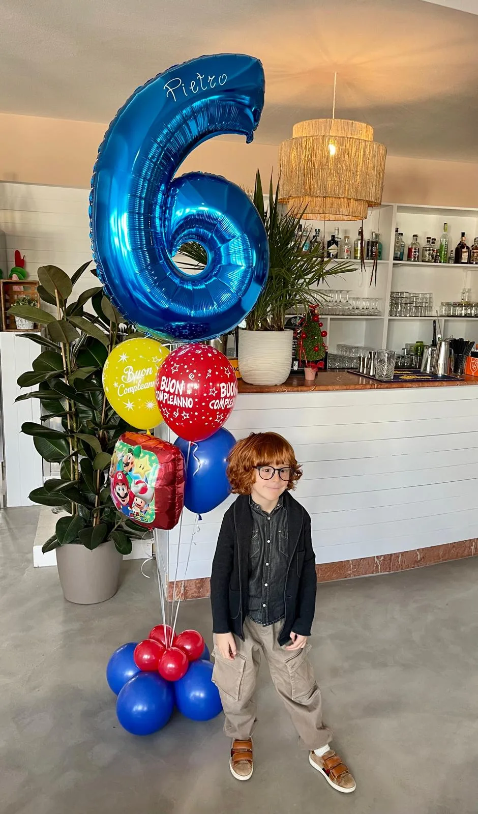 Bambino accanto a un numero 6 blu in mylar con il nome Pietro, palloncini Buon Compleanno rossi e gialli e un palloncino di Super Mario