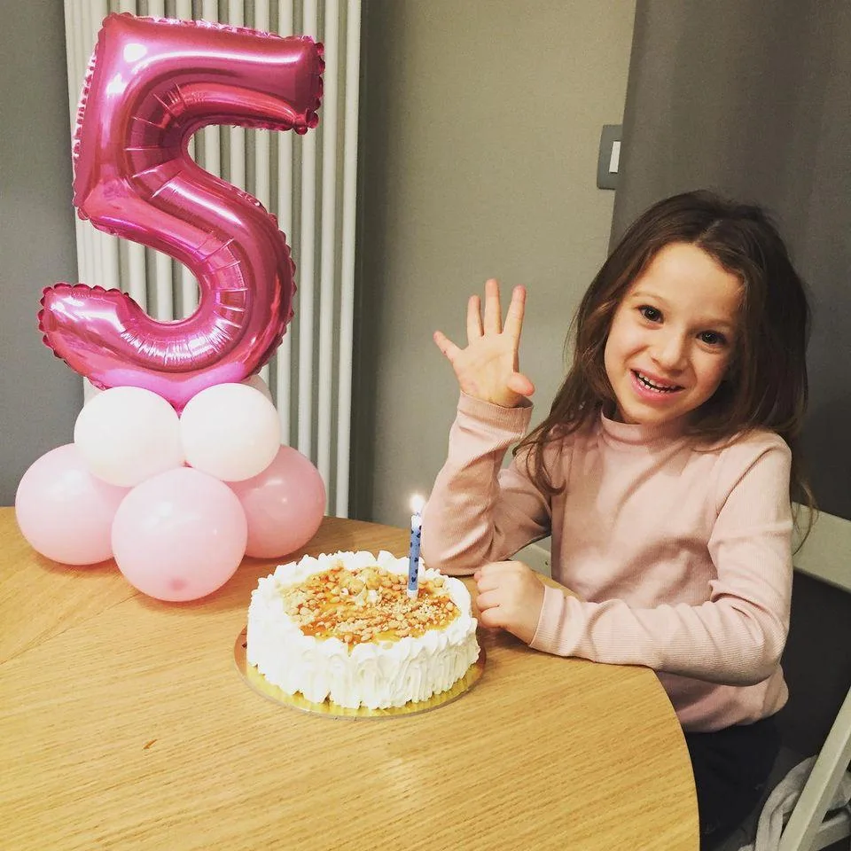 Bambina davanti alla torta con una candelina accesa, accanto a un numero 5 fucsia in mylar su una base di palloncini rosa e bianchi