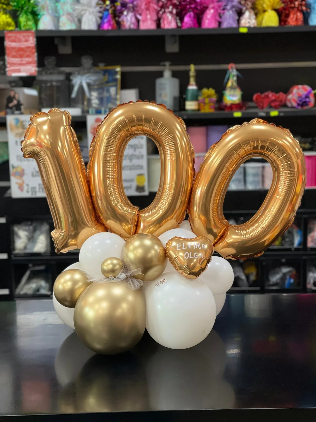 Composizione con il numero 100 in mylar oro su base di palloncini bianchi e oro e un cuore con due nomi scritti a mano, sul bancone del negozio