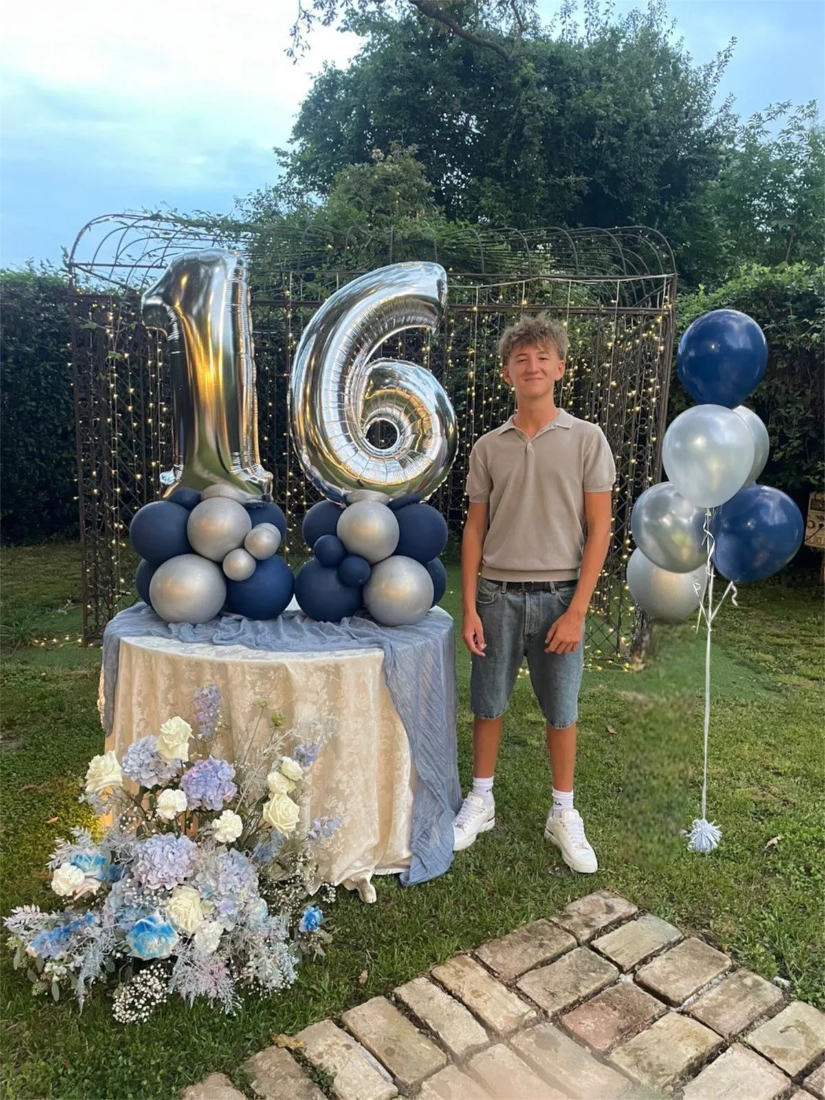 Numero 16 in mylar argento su due basi di palloncini blu e argento, allestito in giardino accanto a un tavolo con fiori