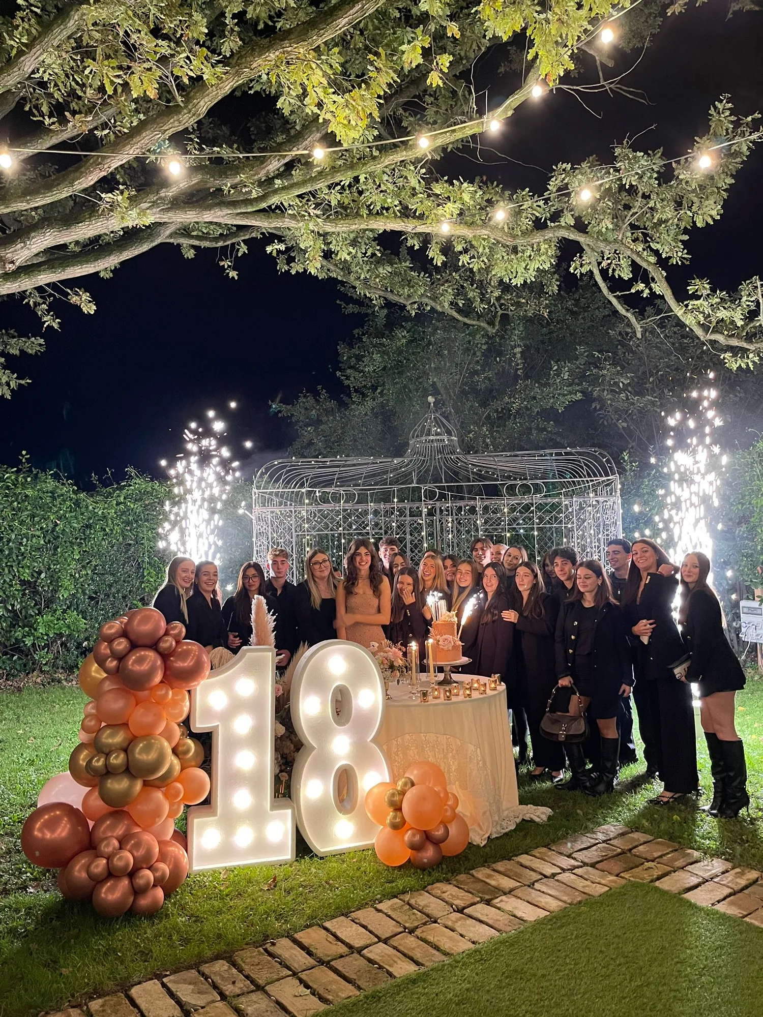 Gruppo di ragazzi attorno al tavolo della torta in giardino di sera, con numero 18 luminoso a lampadine, ghirlanda di palloncini pesca e rame e fontane luminose accese