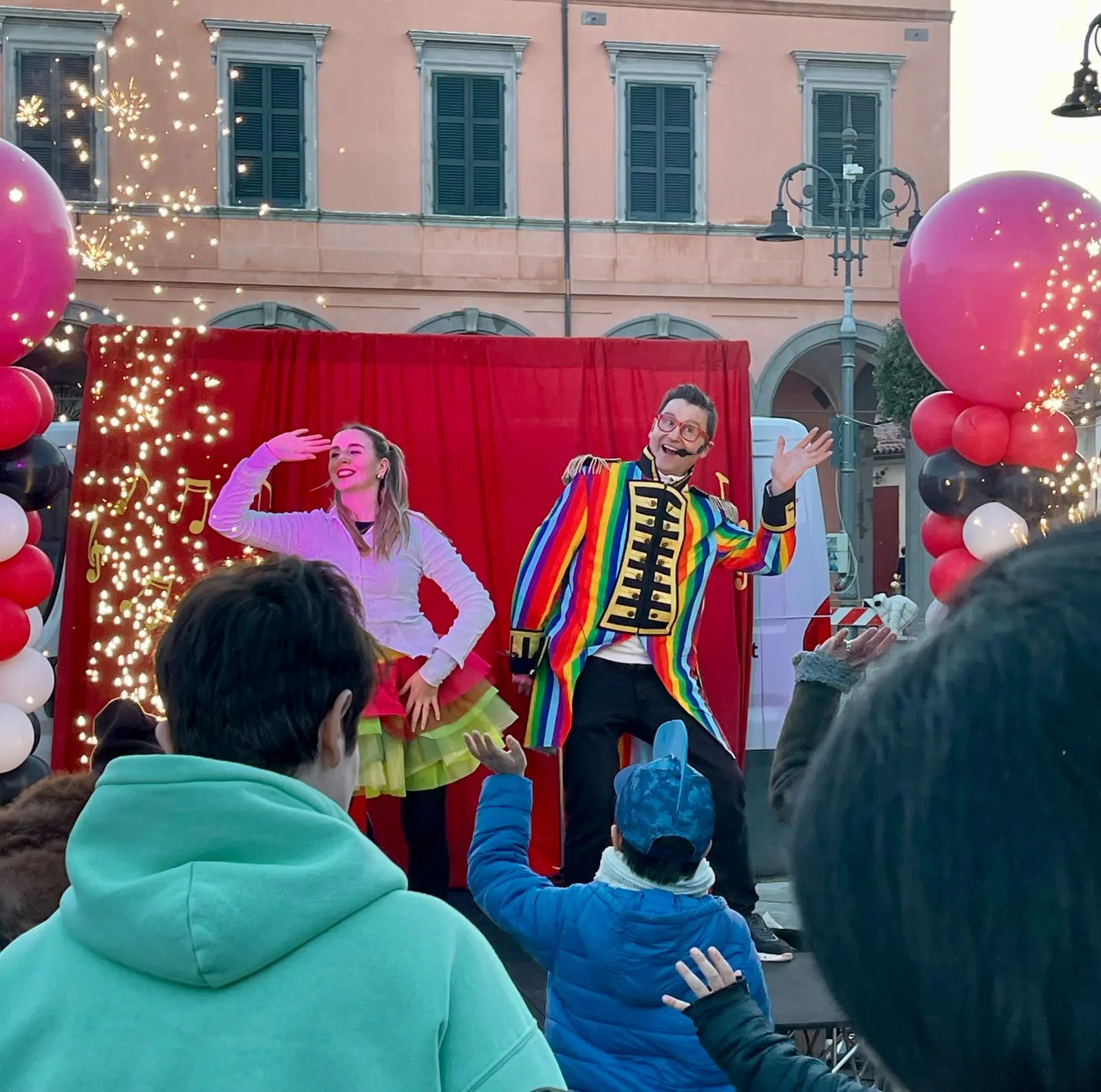 Animatore con giacca arcobaleno e ballerina con tutù colorato sul palco rosso, con i bambini che ballano davanti